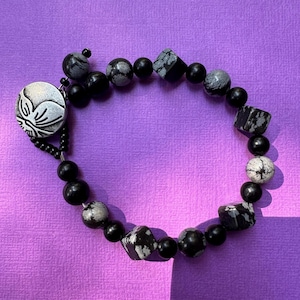 May include: A beaded bracelet featuring a mix of black and gray snowflake obsidian and round black beads. A silver-toned charm with a floral design is attached. The bracelet is displayed on a purple surface.