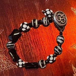 May include: A black and white beaded bracelet with a silver clasp. The bracelet features a variety of beads, including round beads with white polka dots, striped beads, and black rectangular beads. The clasp has an Om symbol.