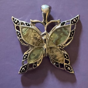 May include: A silver-toned butterfly pendant with iridescent wings and black accents. The butterfly design features a central body and two pairs of wings, with a loop at the top for a chain. The wings have a mother-of-pearl appearance.
