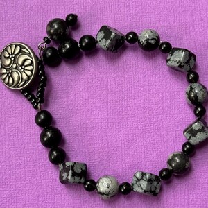 May include: A beaded bracelet featuring black onyx beads and snowflake obsidian stones. The bracelet has a silver-toned button clasp with a floral design. The bracelet is displayed on a purple surface.