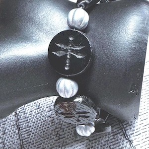 May include: Black beaded bracelet featuring a dragonfly design. The bracelet has a mix of black, silver, and clear beads. The focal point is a black circular bead with a dragonfly etched into it. The bracelet is displayed on a black mannequin.