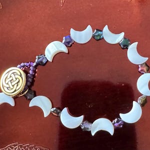 May include: A handmade bracelet featuring crescent moon-shaped beads. The bracelet has a gold-colored clasp with an intricate design and is strung with purple beads and iridescent accents. The bracelet is displayed on a reddish-brown surface.
