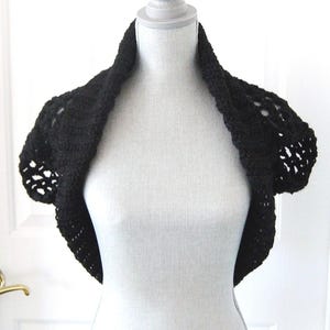 May include: A black, crocheted shrug displayed on a white mannequin. The shrug features a short-sleeved design with openwork detailing on the sleeves. The garment is made of a dark, solid-colored yarn, and the neckline is a rounded collar.