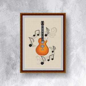 Op de afbeelding: Een ingelijst kruissteekkunstwerk met een oranje en bruine elektrische gitaar omringd door muzieknoten. De gitaar staat centraal, met zwarte noten en gebogen lijnen die een muzikaal thema creëren. Het kunstwerk staat tegen een beige achtergrond.