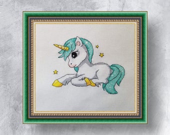 May include: A framed cross-stitch embroidery of a white unicorn with a teal mane and tail. The unicorn is lying down with its head turned to the side. There are small yellow stars in the background.