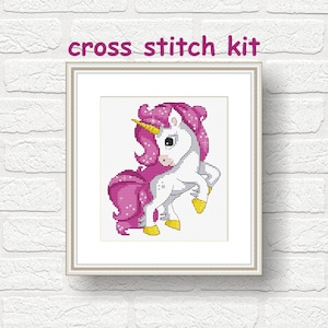 May include: A framed cross stitch kit featuring a cartoon unicorn with a pink mane and tail, a golden horn, and yellow hooves. The text "cross stitch kit" is above the framed image. The background is a white brick wall.
