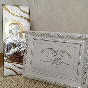 Pode incluir: Dois itens religiosos são exibidos. Um ícone prateado e dourado com a imagem de uma figura religiosa e uma criança. Ao lado, um ponto cruz emoldurado em branco com mãos formando um coração, segurando uma mão menor.