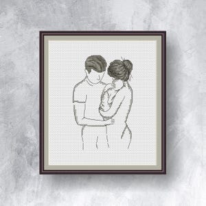 May include: A framed artwork featuring a line drawing of a family. The image depicts a parent holding a baby, with the other parent embracing them. The artwork is in a dark brown frame, set against a textured, light gray background.