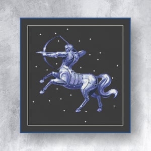 Op de afbeelding: Een ingelijste kruissteekafbeelding van het sterrenbeeld Boogschutter. De afbeelding toont een centaurfiguur met pijl en boog, in blauw en wit tegen een donkergrijze achtergrond met witte steraccenten.