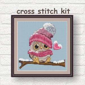 May include: Cross stitch kit featuring a cute cartoon owl wearing a pink hat and scarf, holding a red heart. The owl is sitting on a branch with snow on it.