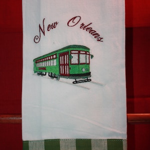 May include: White kitchen towel with a green and white checkered border. The towel has red embroidered text that says "New Orleans" and an embroidered image of a green streetcar.