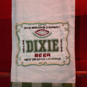 May include: White kitchen towel with a green and white checkered border. The towel features an embroidered logo for Dixie Brewing Company, New Orleans, Louisiana. The logo is in a gold and green frame with the words "Dixie Beer" in red.