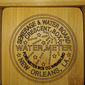 May include: A close-up of a wooden coaster with a black and white engraved design. The design features the text "SEWERAGE & WATER BOARD CRESCENT BOX WATER METER FORD METER BOX CO. WABASH.IND. NEW ORLEANS, LA."