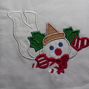 May include: Embroidered white kitchen towel with a snowman design. The snowman is wearing a brown hat and a red bow. The snowman is holding a candy cane and is surrounded by holly leaves.