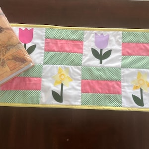 May include: A yellow, green, and pink patchwork table runner with a checkered pattern and appliqued flowers. The flowers are pink, purple, and yellow.