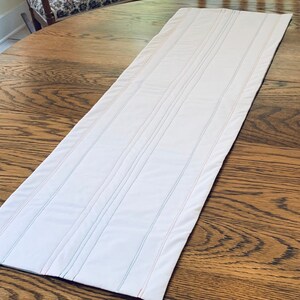 May include: White table runner with thin, colorful stripes. The runner is quilted with straight lines and has a slightly textured surface.
