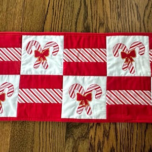 May include: A red and white Christmas table runner with a candy cane design. The runner features alternating squares of red and white striped fabric, and squares with candy cane appliqués with red bows. A festive holiday decoration.