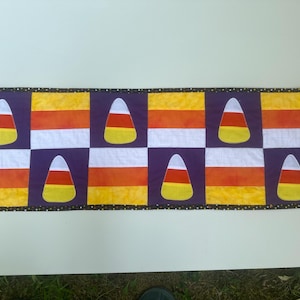 May include: A purple, yellow, orange, and white fabric table runner with a Halloween candy corn theme. The runner has a black border with white polka dots.