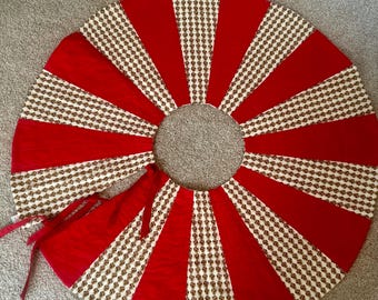 May include: A round Christmas tree skirt with a red and cream color scheme. The skirt is divided into alternating red velvet and cream-colored sections with a repeating gold pattern. The skirt has a tie closure.