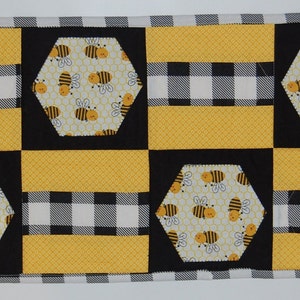 May include: A yellow and black table runner with a honeycomb pattern and bees. The runner is made of fabric and has a checkered pattern.