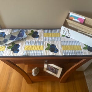 May include: A white table runner with a blue and white gingham pattern and yellow stripes. The runner is decorated with blue and green blueberry appliques. The runner is on a wooden table with a white organizer that says "ORGANIZE" on the front.
