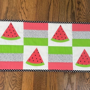 May include: A watermelon-themed table runner with a black and white polka dot border. The runner is made of fabric squares with alternating patterns of green, pink, and white. Each square features a watermelon slice with black seeds.
