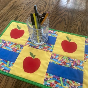 May include: A yellow and blue quilted table runner with a green border. The runner has a repeating pattern of red apples and colorful pencils. A glass jar with black and yellow pens and pencils sits on top of the runner.