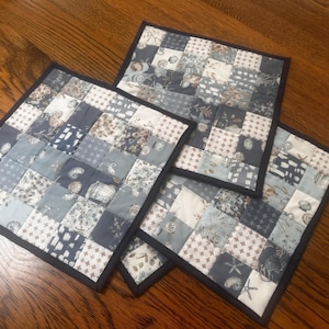 May include: Three square quilted placemats with a patchwork design. The squares feature various patterns in shades of blue, gray, and white, with a black border. The placemats are arranged on a wooden surface.