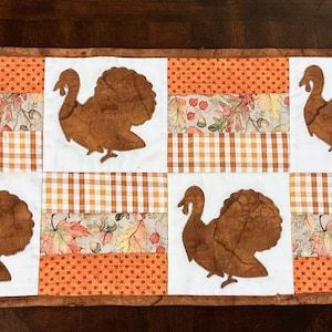 May include: A brown and white quilted table runner with a Thanksgiving theme. The runner features six brown turkey silhouettes on a white background. The runner is made of fabric with a brown and white checkered pattern, a brown and orange floral pattern, and a solid orange pattern.