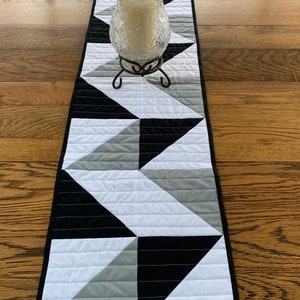 May include: A black, white, and gray geometric table runner with a candle holder in the center.