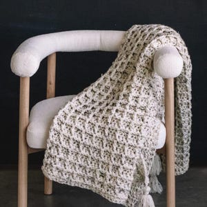 May include: A cream-coloured, chunky knit throw blanket with a waffle-like texture and long, matching tassels at the corners, draped over a light-toned wooden chair with a curved backrest and upholstered seat in a matching cream fabric.