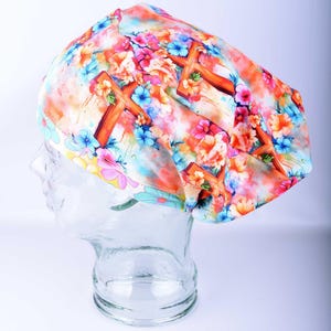 A full-coverage style medical scrub cap featuring a colorful watercolor pattern of Christian crosses in soft pastel shades of pink, blue, orange, and purple on a white background. The cap is displayed on a clear glass mannequin head.