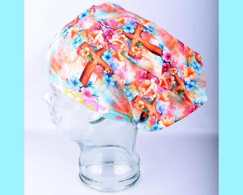 A full-coverage style medical scrub cap featuring a colorful watercolor pattern of Christian crosses in soft pastel shades of pink, blue, orange, and purple on a white background. The cap is displayed on a clear glass mannequin head.