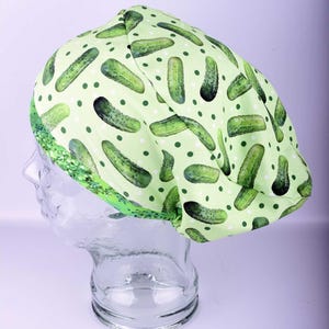May include: A light green surgical scrub cap featuring a pattern of green pickles and small dark green polka dots. The cap has a green and white patterned border. The cap is displayed on a clear mannequin head.