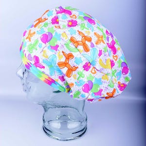 May include: A white scrub cap with a colorful pattern of balloon animals, flowers, and stars. The cap has a tie-dye headband and is displayed on a clear mannequin head. The design includes orange, pink, blue, green, and yellow elements.