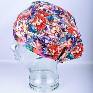 May include: A colorful, patterned hat featuring cartoon illustrations of princesses. The hat is displayed on a clear mannequin head. The design includes shades of red, blue, purple, and gold, creating a vibrant and eye-catching accessory.