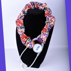 May include: A stethoscope with a decorative cover featuring a colorful pattern of figures in red, purple, and blue. The stethoscope is displayed on a black mannequin head.
