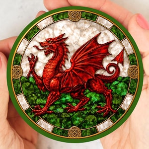 May include: Round coaster featuring a red dragon with outstretched wings, a symbol of Wales. The dragon is set against a green and white background, framed by a green border with gold Celtic knot accents. The coaster is held by a person.