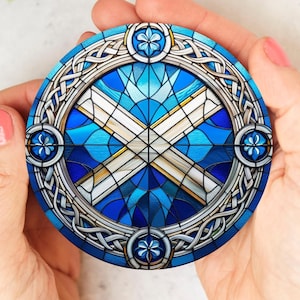 Peut inclure: Sous-verre rond avec un motif de vitrail représentant le drapeau écossais, une croix de Saint-André blanche sur fond bleu. Le dessous de verre a une bordure décorative argentée avec des nœuds celtiques et des motifs floraux. Le dessous de verre est tenu dans deux mains.