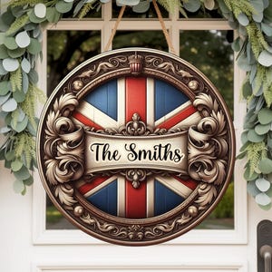 Op de afbeelding: Een rond houten bord met het ontwerp van de Union Jack-vlag en de tekst "The Smiths" in een banner. Het bord heeft een sierlijke, decoratieve rand en wordt opgehangen met een touw. De achtergrond omvat een witte deur en groen.