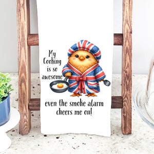 May include: A white kitchen towel with a cartoon illustration of a chick wearing a chef's hat and a robe with a Union Jack pattern. The text on the towel reads "My Cooking is so awesome even the smoke alarm cheers me on!"
