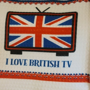 May include: A white waffle-weave kitchen towel featuring a graphic of a television set displaying the Union Jack flag. Below the television, the text reads I LOVE BRITISH TV in blue letters. Decorative borders of small Union Jack flags are present.