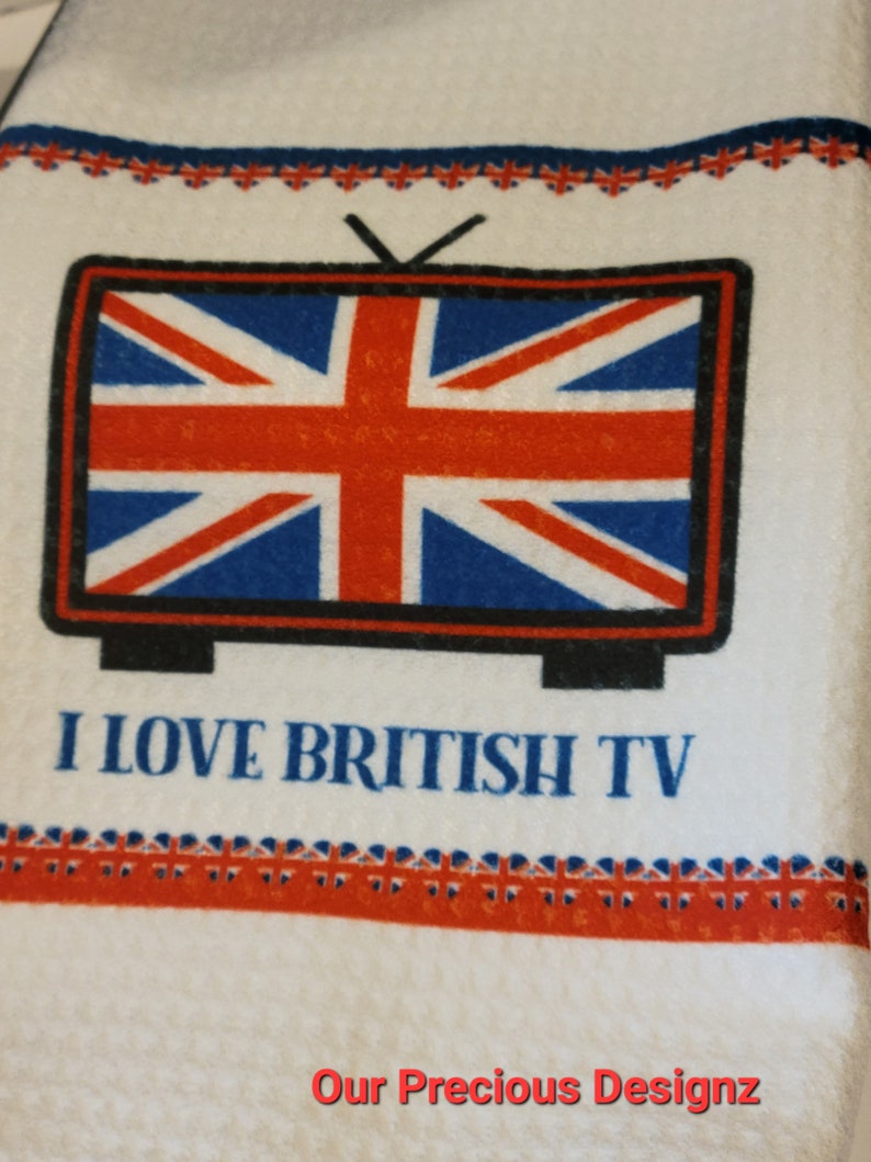 May include: A white waffle-weave kitchen towel featuring a graphic of a television set displaying the Union Jack flag. Below the television, the text reads I LOVE BRITISH TV in blue letters. Decorative borders of small Union Jack flags are present.