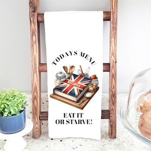 Puede incluir: Paño de cocina blanco con el texto "TODAYS MENU" y "EAT IT OR STARVE!". El paño presenta un libro de cocina con un diseño de la Union Jack, rodeado de utensilios e ingredientes de cocina. El paño está colgado de una escalera de madera.