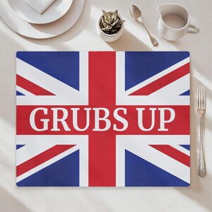 Könnte beinhalten: Ein rechteckiger Tischset mit dem Union Jack-Flaggen-Design in Rot, Weiß und Blau. Die Worte "GRUBS UP" sind weiß in der Mitte aufgedruckt. Das Tischset liegt auf einer weißen Oberfläche mit einer Gabel, einem Löffel und einer kleinen Pflanze.