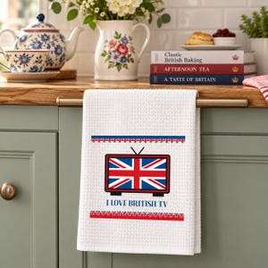 May include: A white waffle-weave kitchen towel featuring a graphic of a television set displaying the Union Jack flag. Below the television, the text reads I LOVE BRITISH TV in blue letters, framed by two horizontal rows of small red and blue flag icons.
