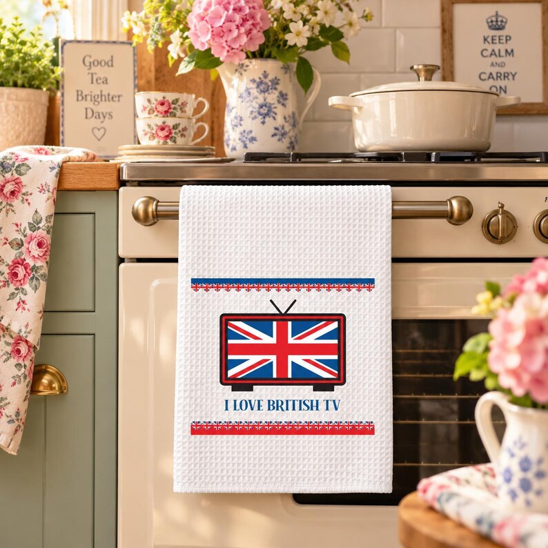 May include: A white waffle-weave kitchen towel featuring a graphic of a television set displaying the Union Jack flag. Below the television, the text reads I LOVE BRITISH TV in blue capital letters, framed by red and blue decorative borders.