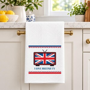 May include: A white waffle-weave kitchen towel featuring a graphic of a television set with a Union Jack flag screen. Above and below the television are decorative borders of small Union Jack flags. The text I LOVE BRITISH TV is printed below.