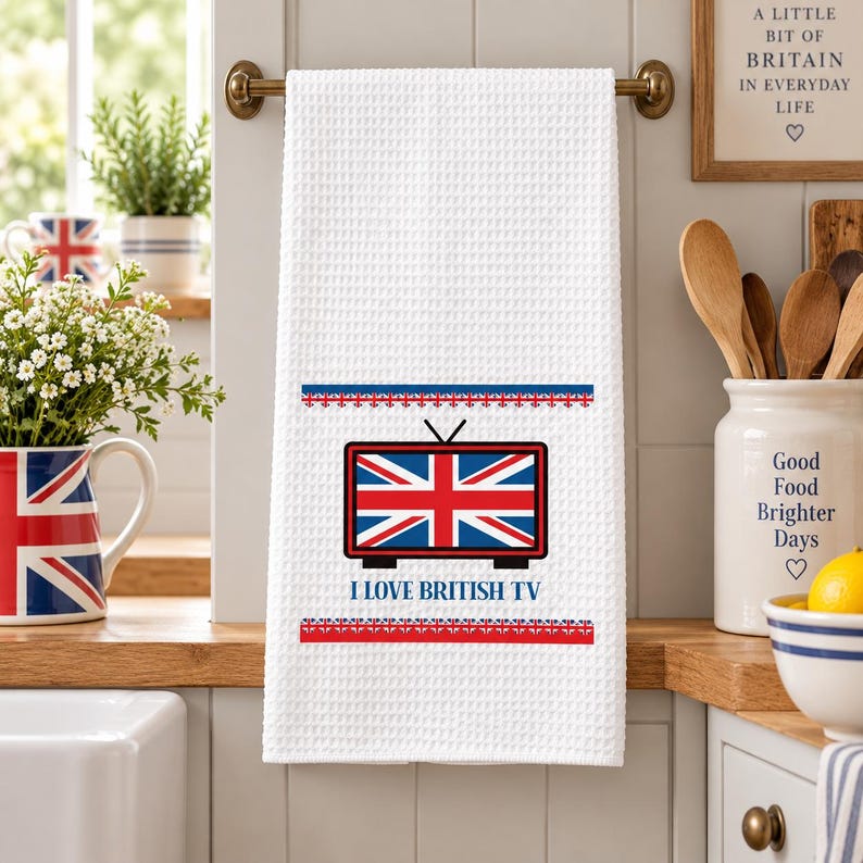 May include: A white waffle-textured kitchen towel hanging on a rod. The towel features a graphic of a television set displaying the Union Jack flag, with the text I LOVE BRITISH TV printed below it, framed by two horizontal rows of small flag icons.