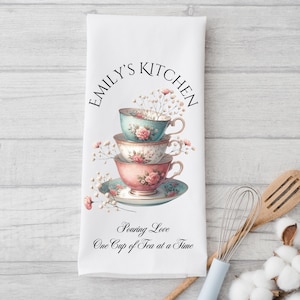 May include: White kitchen towel with a floral design featuring three stacked teacups. The text "Emily's Kitchen" is printed in a circle at the top of the towel. The text "Pouring Love One Cup of Tea at a Time" is printed at the bottom of the towel.